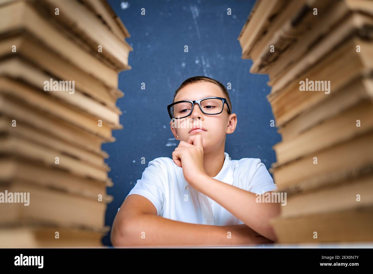 Smart boy in glasses sitting between two piles of books and look down