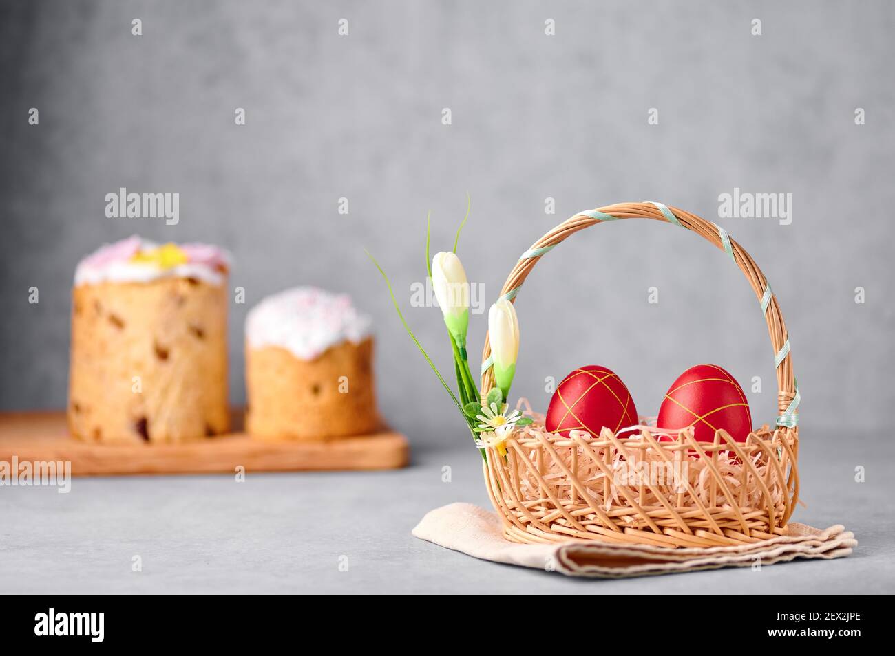 Two red painted Easter eggs in the wicker basket on gray table top with two easter cakes on backdrop Stock Photo