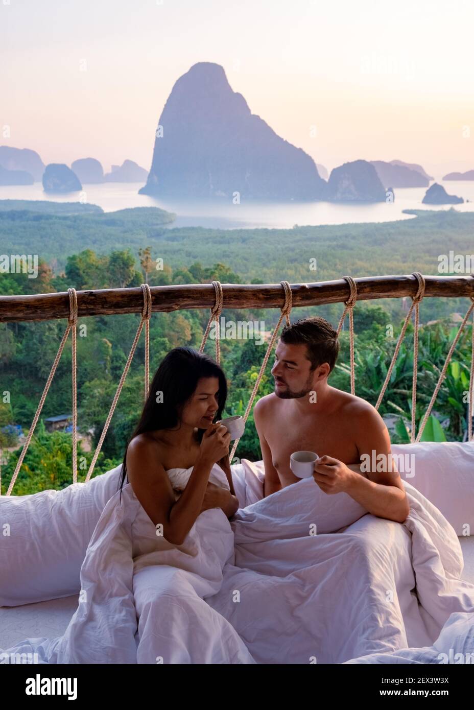 Phangnga Bay, couple waking up in bed in nature jungle looking out over ocean and jungle during sunrise at the wooden hut in the mountains of Thailand men and woman mid age morning sunrise Phanga Thailand Stock Photo