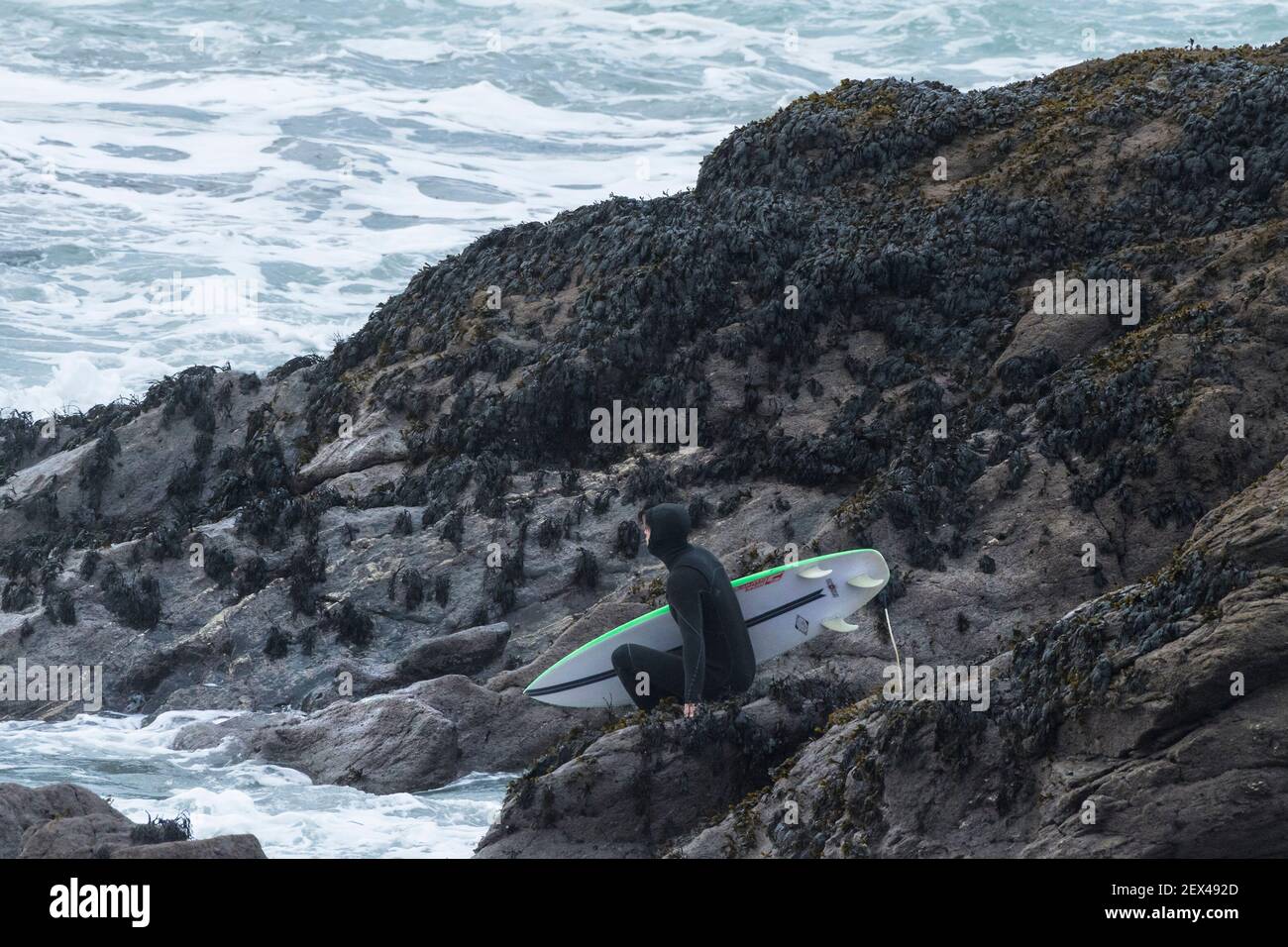A surfer holding his surfboard sitting on rocks and waiting for the right moment to enter the sea at the rugged Little Fistral in Newquay in Cornwall. Stock Photo