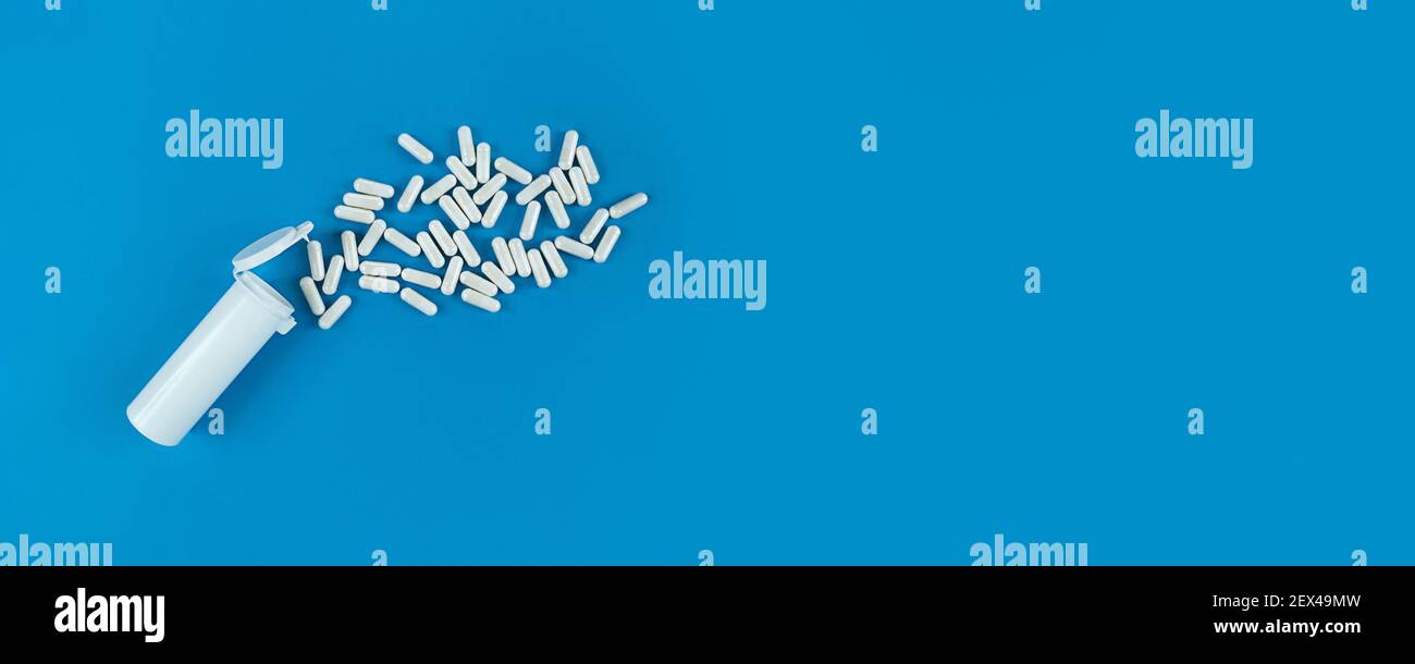 White plastik medical bottle and scattered pills capsules on blue backdrop. Banner with copy space. Stock Photo