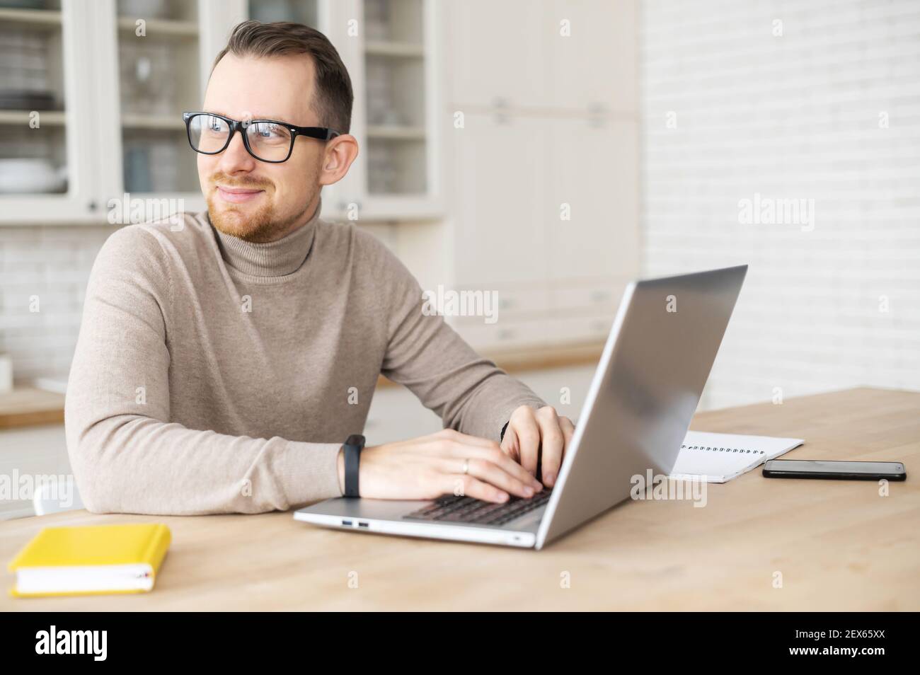 Handsome young hipster freelancer man with beard in glasses sitting at the table and looking away, taking a break from working on laptop, dreaming about vacation or salary, enjoying working from home Stock Photo