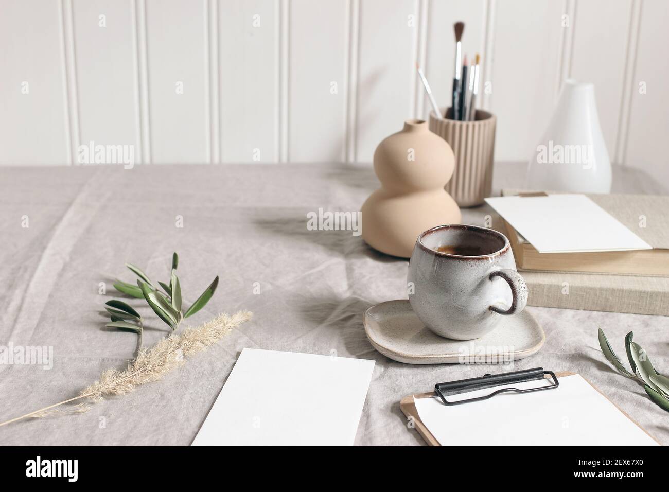 Artistic wokspace, still life. Paint brushes and pencils. Olive branches, vase, cup of coffee and blank paper card mockups on linen tablecloth. Art Stock Photo
