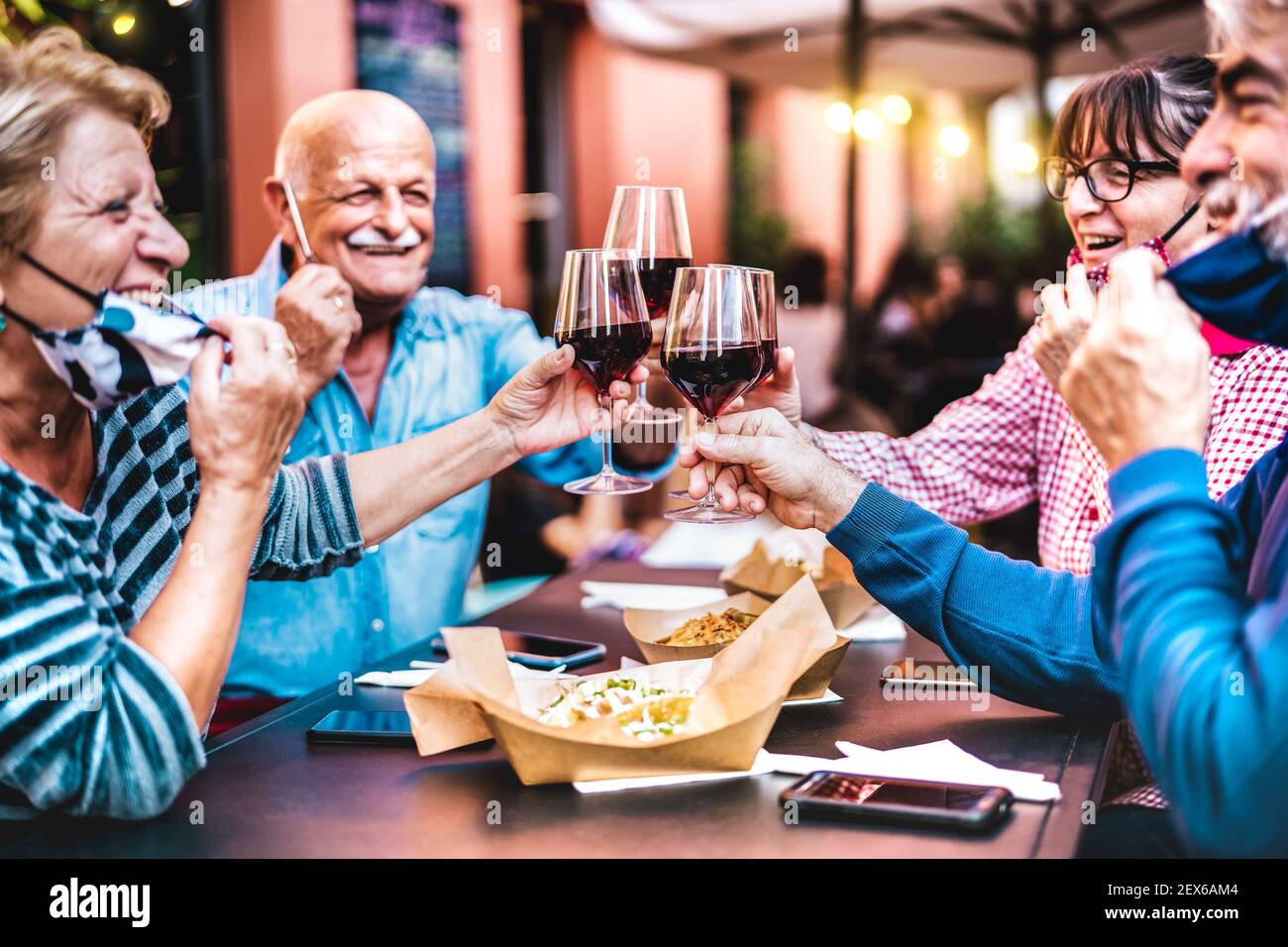 Senior people toasting wine at restaurant bar wearing open face masks - New normal life style concept with happy mature friends having fun together Stock Photo