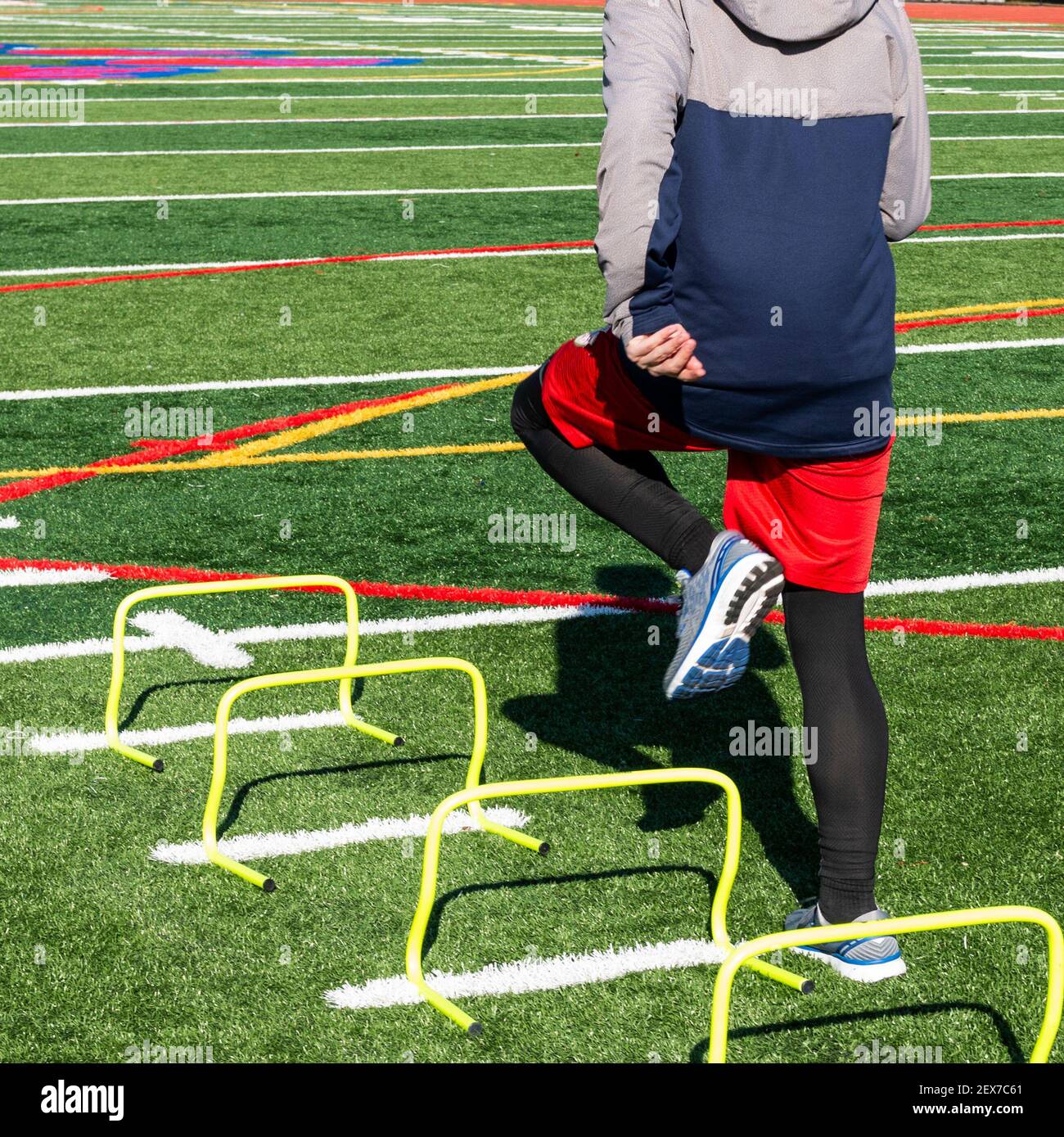 Rear view of a high school track runner is stepping over one foot mini hurdles on a turf field on a cold spring day at track and field practice. Stock Photo