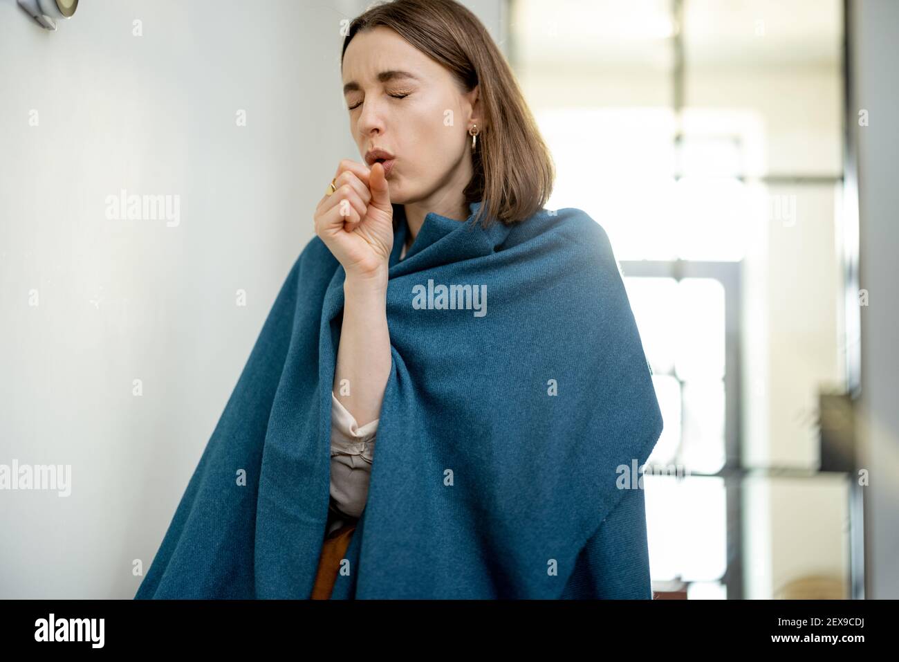 Pretty woman is coughs and cold, covered with a plaid standing near the heat regulator on the wall. Smart home technology is not working. Saving electricity.  Stock Photo