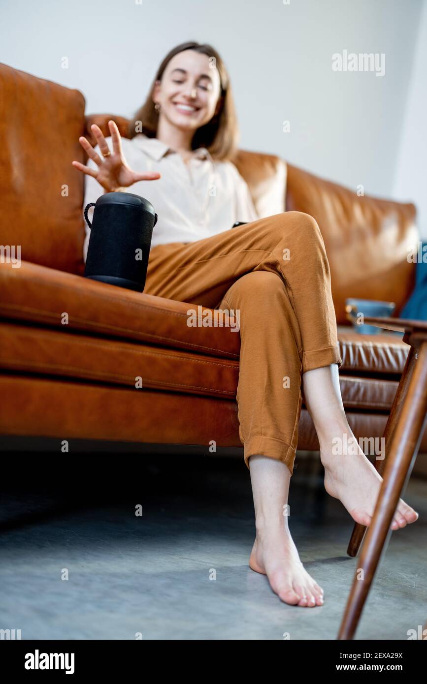 Happy pretty woman speaking with black audio assistant column on the coffee table while sitting on brown leather sofa in stylish living room. Smart home concept. Stock Photo