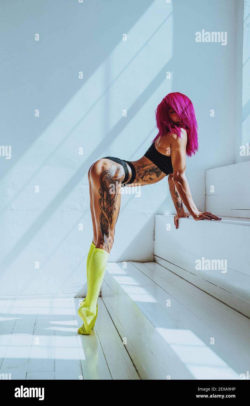 Young sexy slim woman with pink hair standing in bright white interior. Tattoo on body. Stock Photo