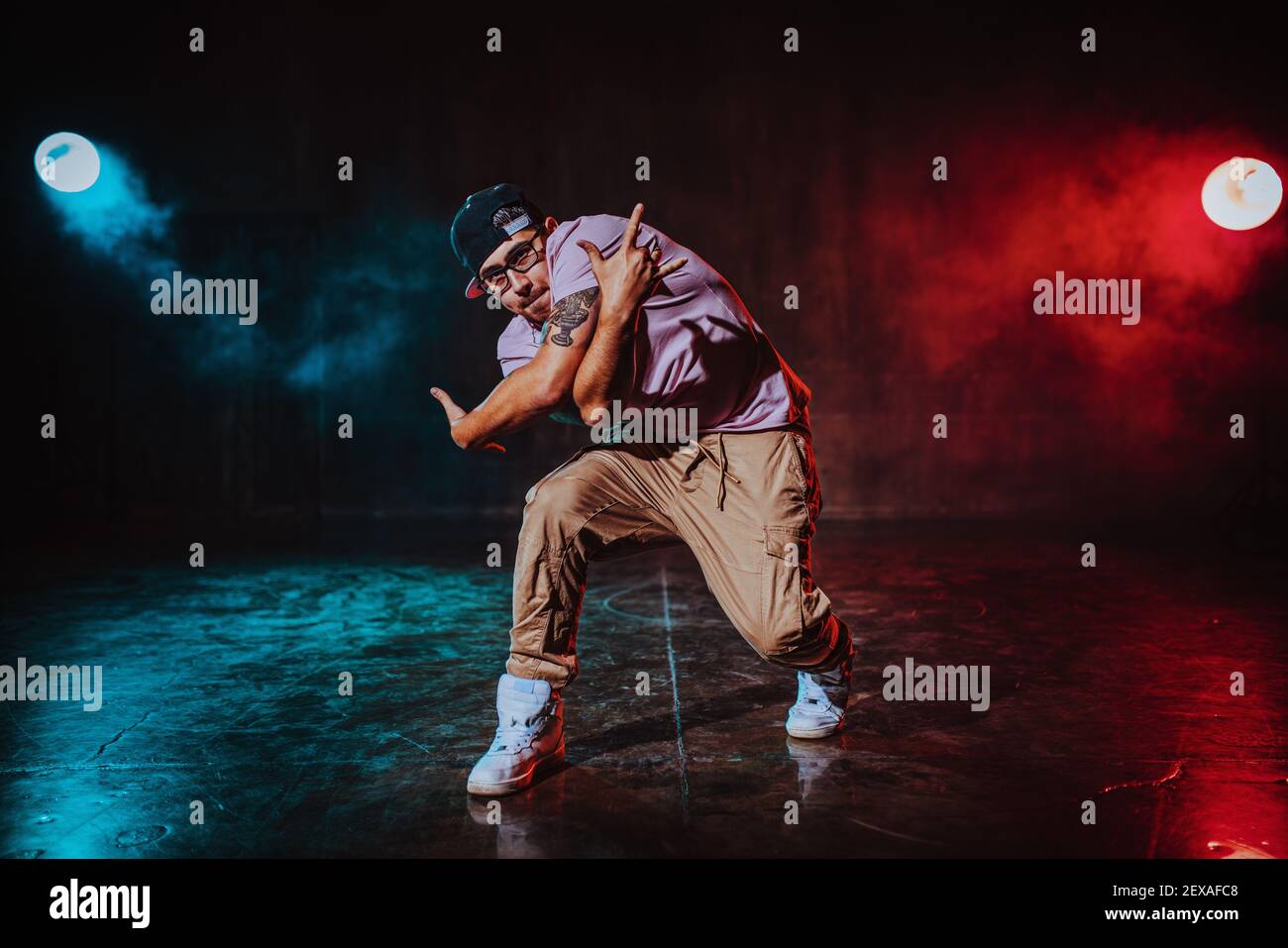Young man break dancing in dark hall with blue and red lights. Tattoo on hand. Stock Photo