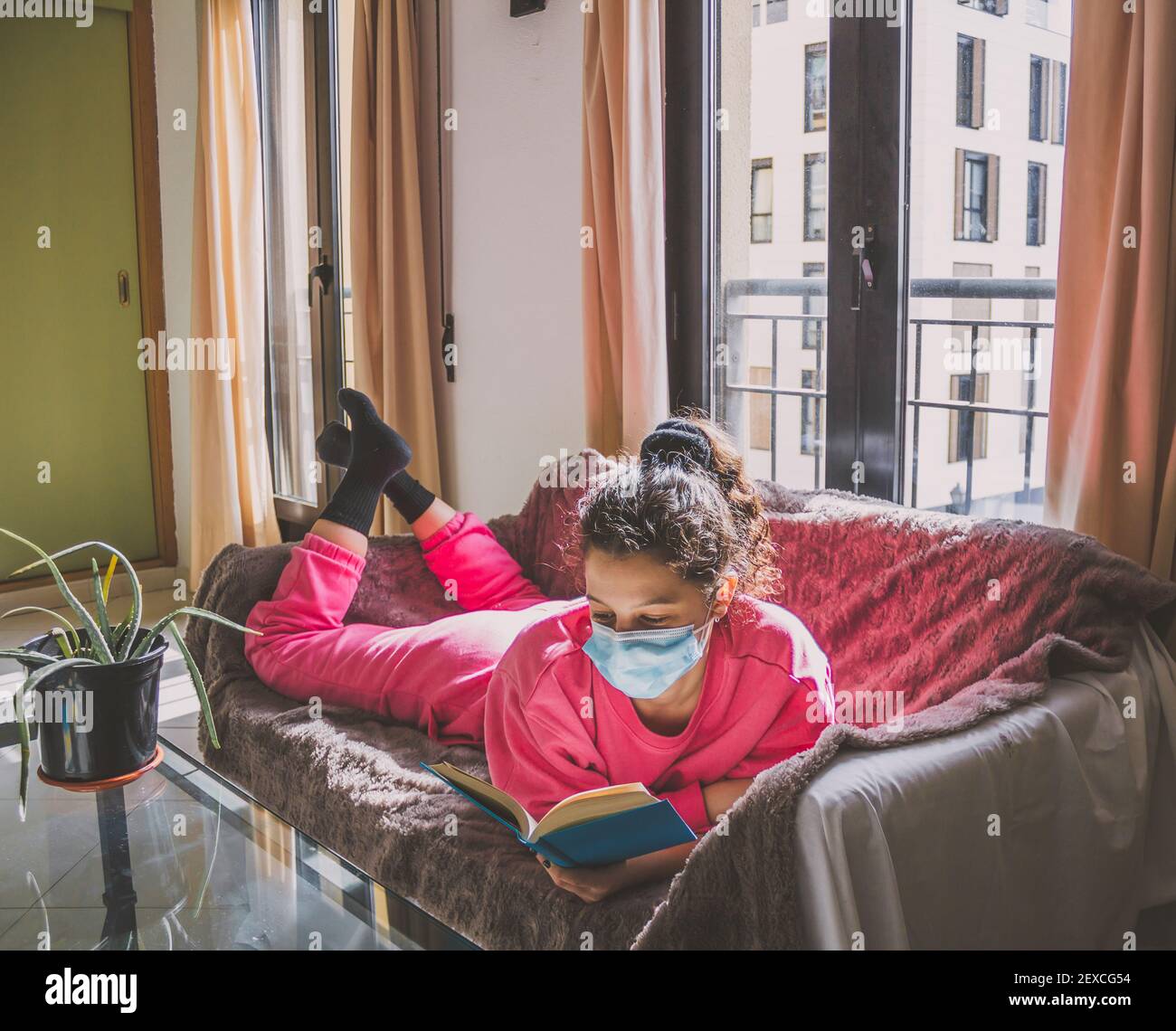 young woman with protective mask lying on the sofa reading a book Stock Photo