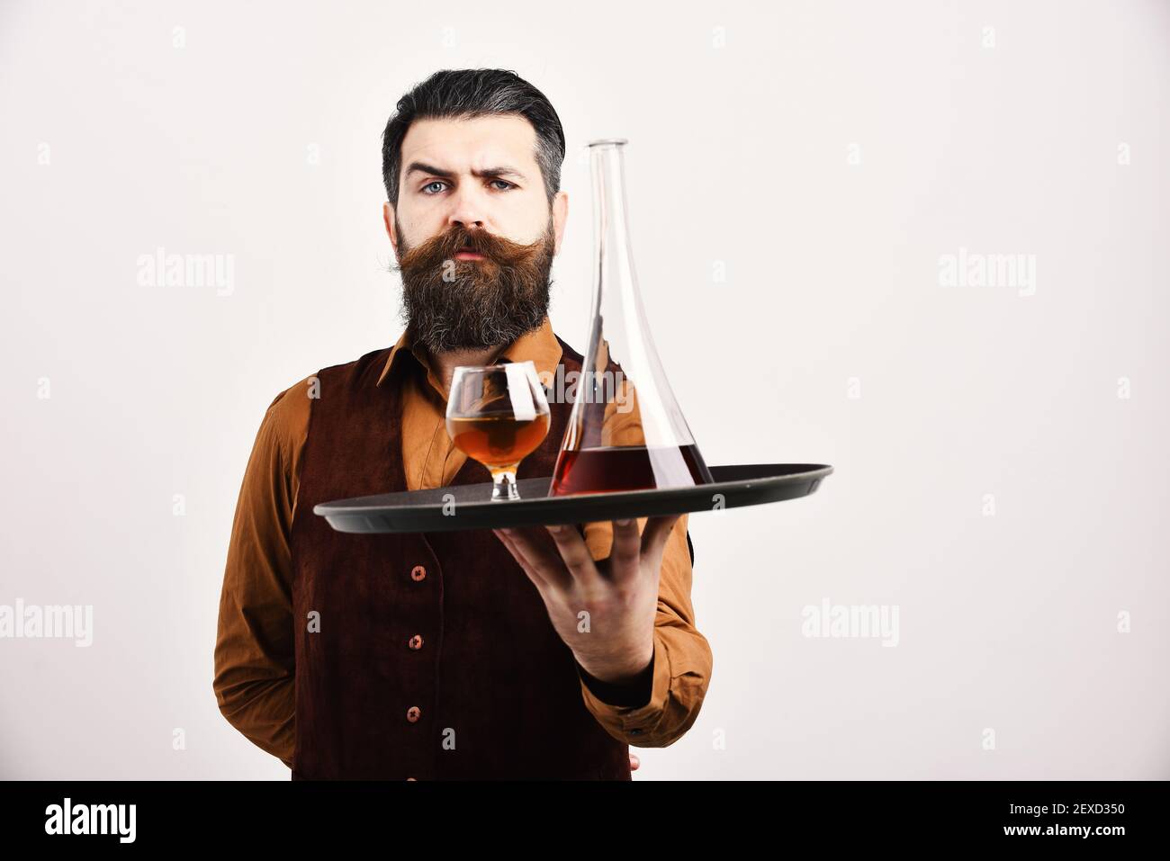 Barman with serious face serves scotch or brandy. Waiter with glass and bottle of whiskey on tray. Man with beard holds cognac on white background, copy space. Restaurant service and drinks concept. Stock Photo