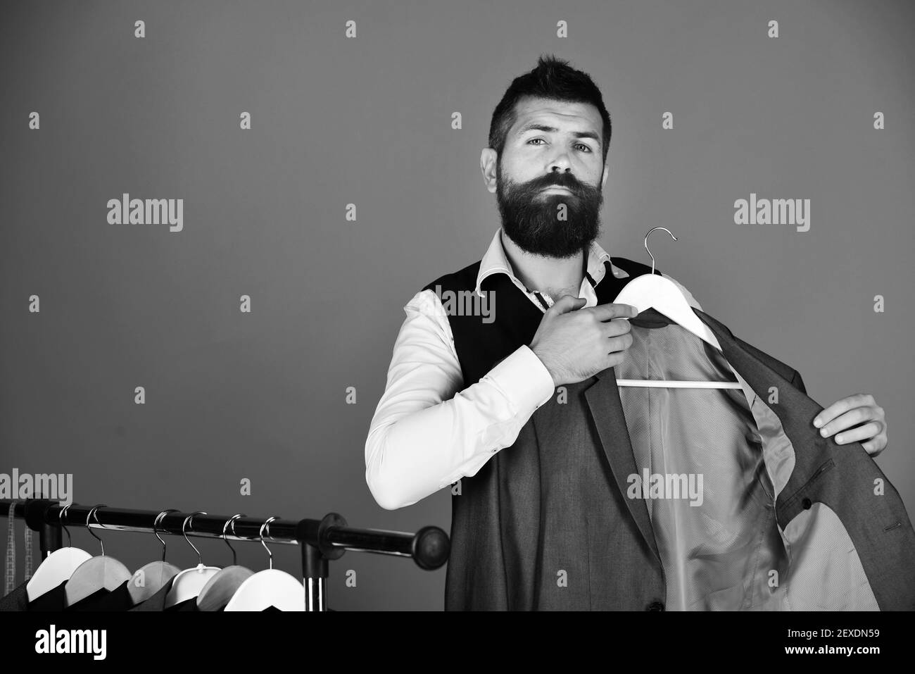 Man with beard in vest by clothes rack. Businessman with proud face near jackets on red background. Official wardrobe choice concept. Shop assistant or seller puts grey suit on clothes hanger Stock Photo