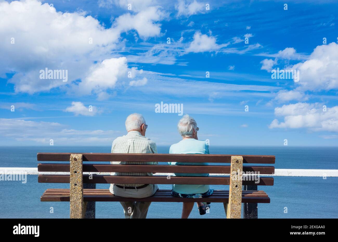 Rear view of elderly couple sitting on seat overlooking the sea on a sunny day. Stock Photo