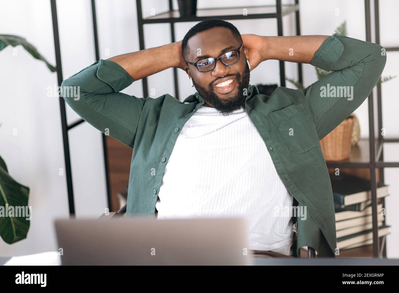 Satisfied freelancer guy relaxing at the workplace and smiling. Confident African American male entrepreneur sits at home office, taking break from online work and dreaming about vacation Stock Photo