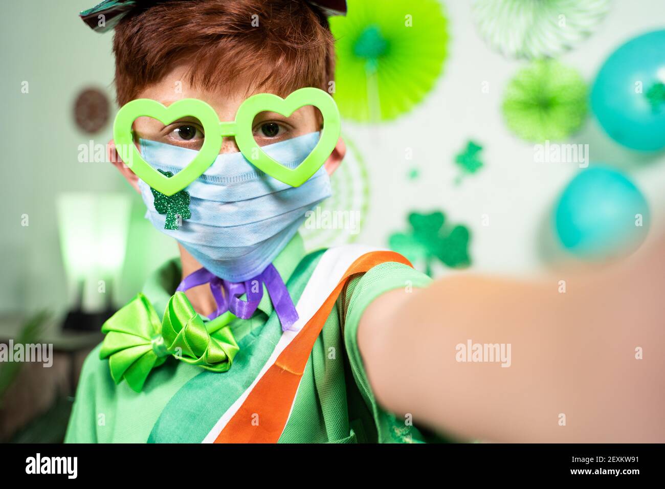 Selective focus on mask, Young kid with medical face mask taking selfie during Saint patricks day celebration at home on decorated background during Stock Photo