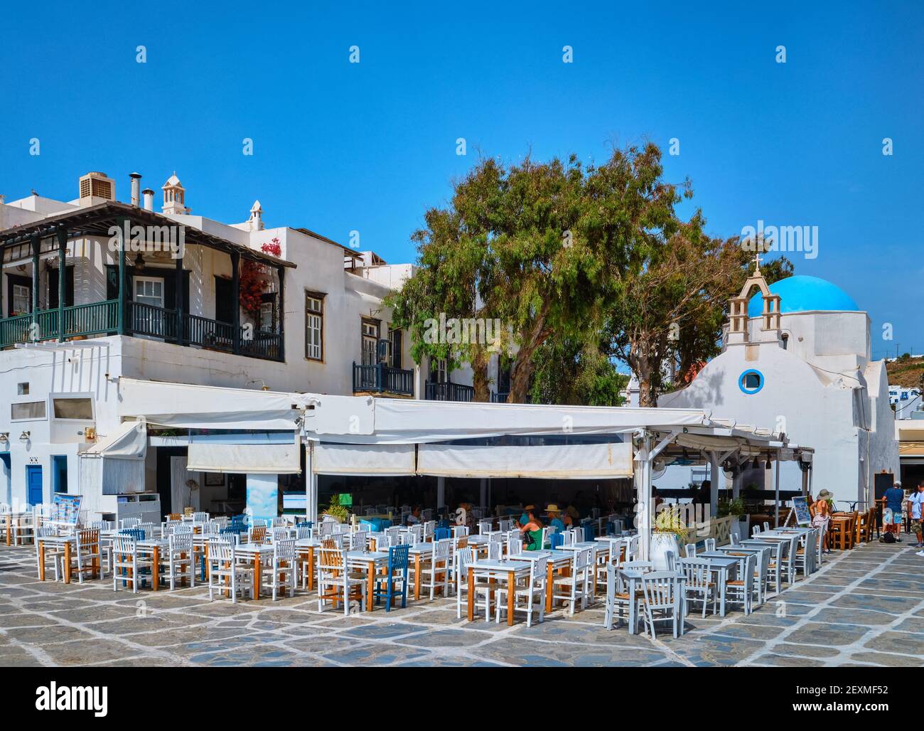 Colorful outdoor cafe or restaurant in typical Greek island town. Greek Orthodox church, blue dome, tourists roam around. Mykonos, Cyclades, Greece. Stock Photo