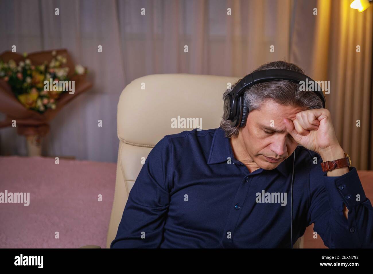 50-year-old man listens to music on headphones at home, sitting in a chair. Relaxing delight. Stock Photo