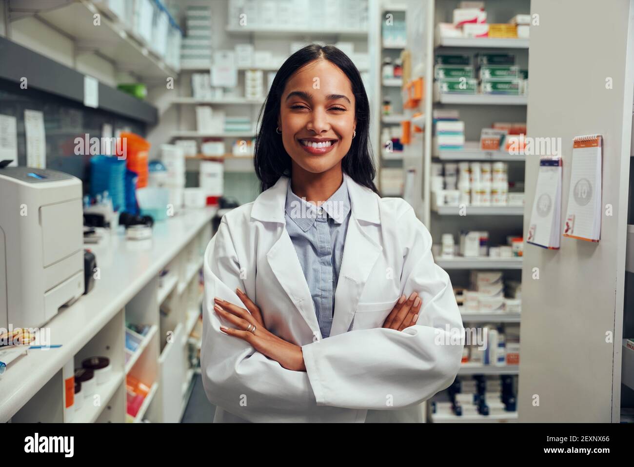 Cheerful young african american female pharmacist wearing labcoat with folded hands standing in chemist near shelves Stock Photo