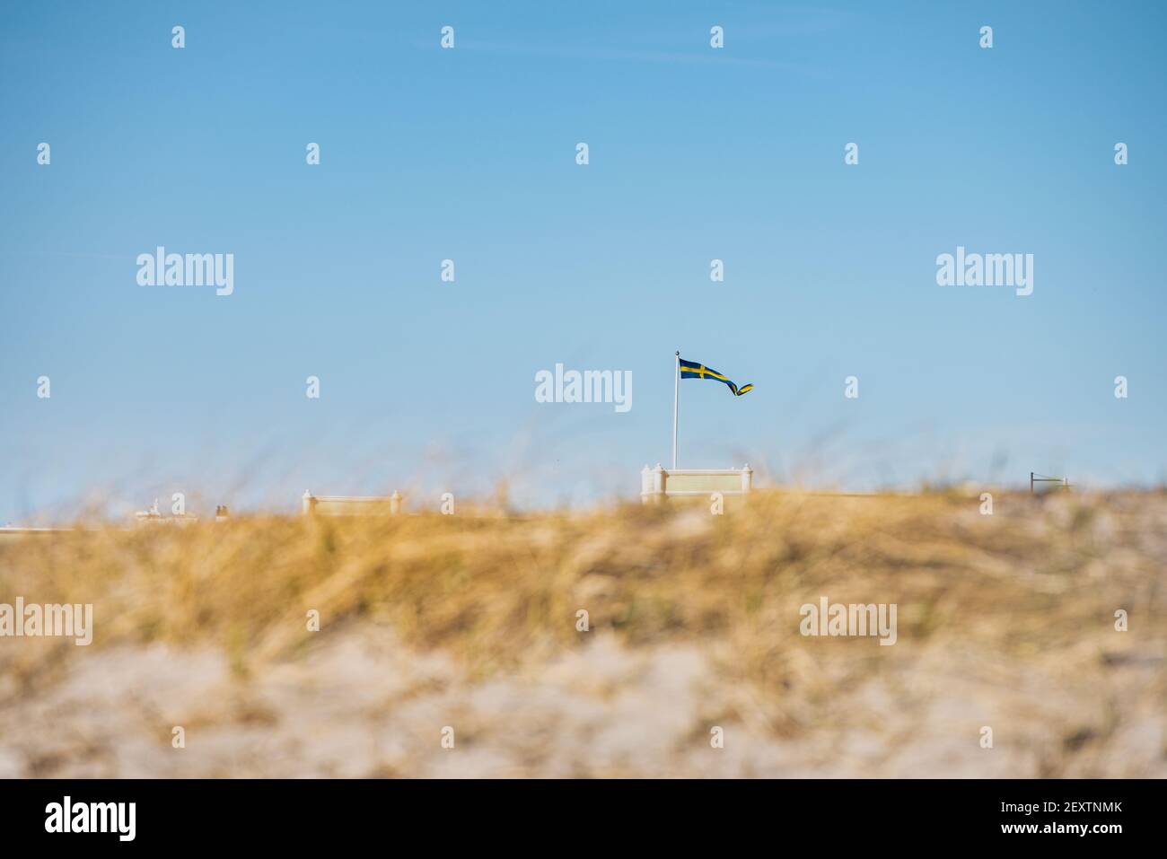 Swedish flag fluttering on a pole, captured through the sandy dunes of a beach. Scandinavian Swedish banderole waving on Ribersborgs Kallbadhus. Malmo Stock Photo