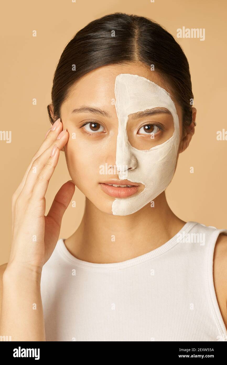 Portrait of young woman with facial mask applied on half of her face receiving spa treatments, posing isolated over beige background. Front view Stock Photo