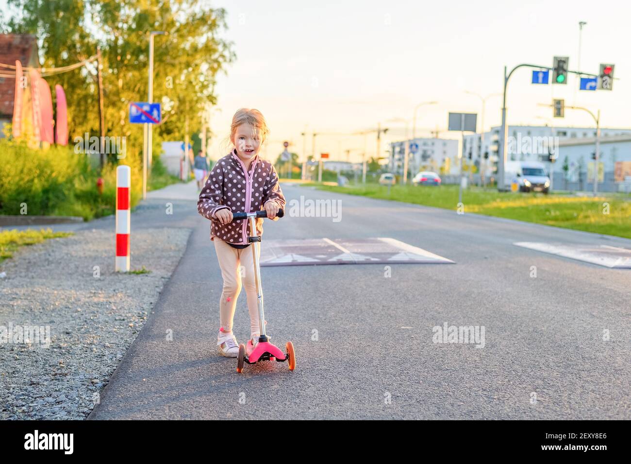 Preschooler girl riding scooter outdoors. Outdoor portrait of cute blonde hair little girl on kick scooter on a road with sunny blurred background Stock Photo