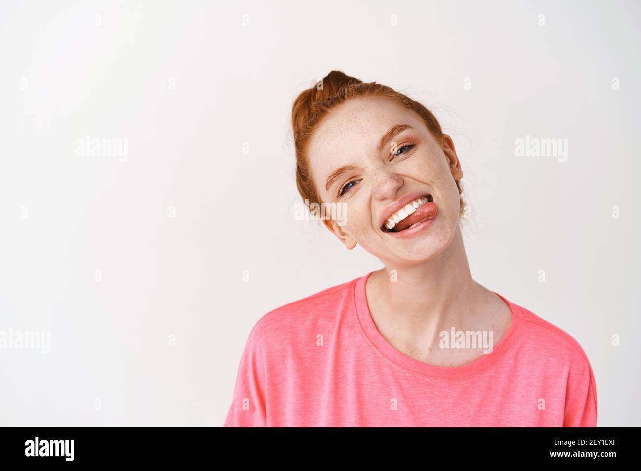 Cheerful teenage girl with ginger hair combed in messy bun, showing white smile and tongue, standing against white background in pink t-shirt Stock Photo