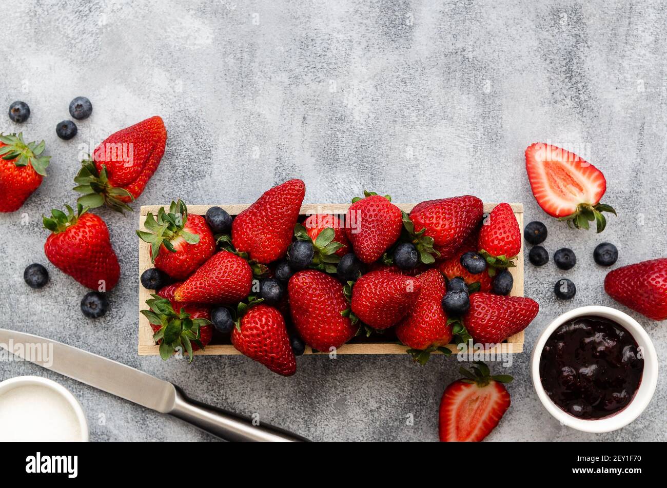 Strawberries and blueberries in a wooden rectangular box, strawberry and blueberry jam and sugar in white bowls with a knife, on grey backdrop. Stock Photo