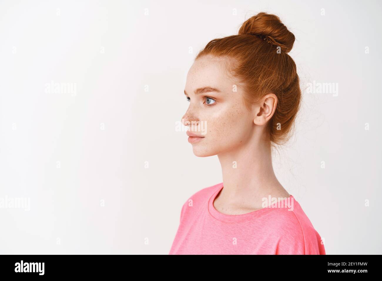 Skincare and makeup concept. Profile of young woman with natural red hair combed in messy bun, looking left, standing over white background. Facial Stock Photo