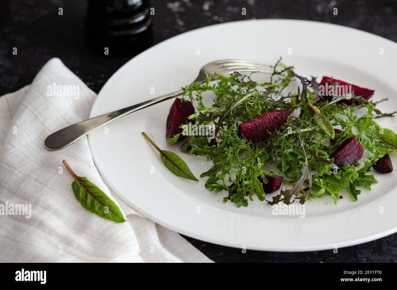 Green and purple mizuna and beetroot salad in a white plate, with a fork and a white napkin, on black backdrop. Stock Photo