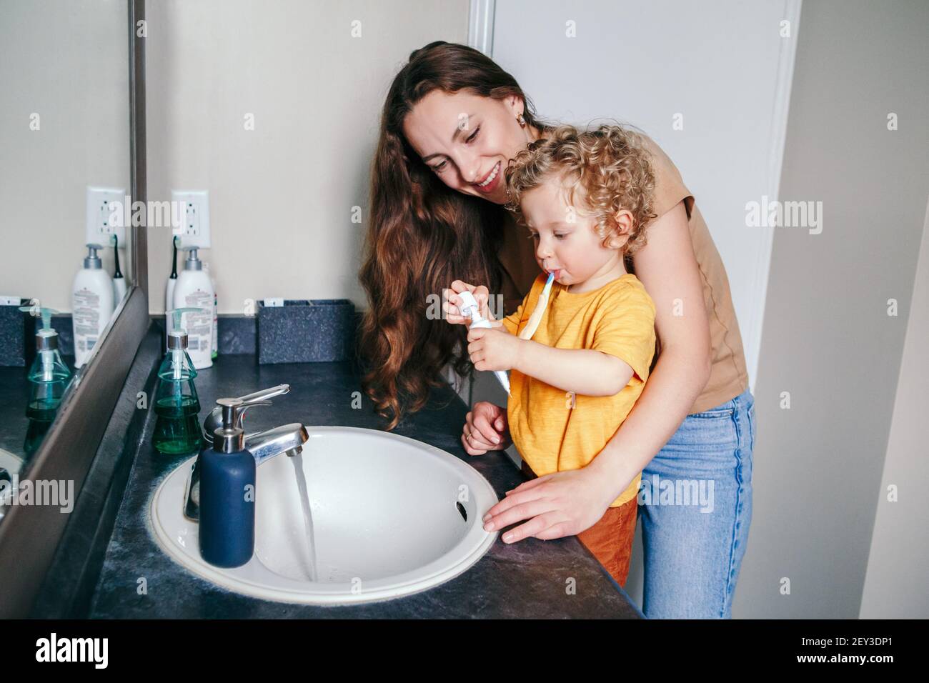 Young Caucasian mother helping boy toddler brush teeth in bathroom at home. Health hygiene and morning routine for children. Mom helping supporting Stock Photo