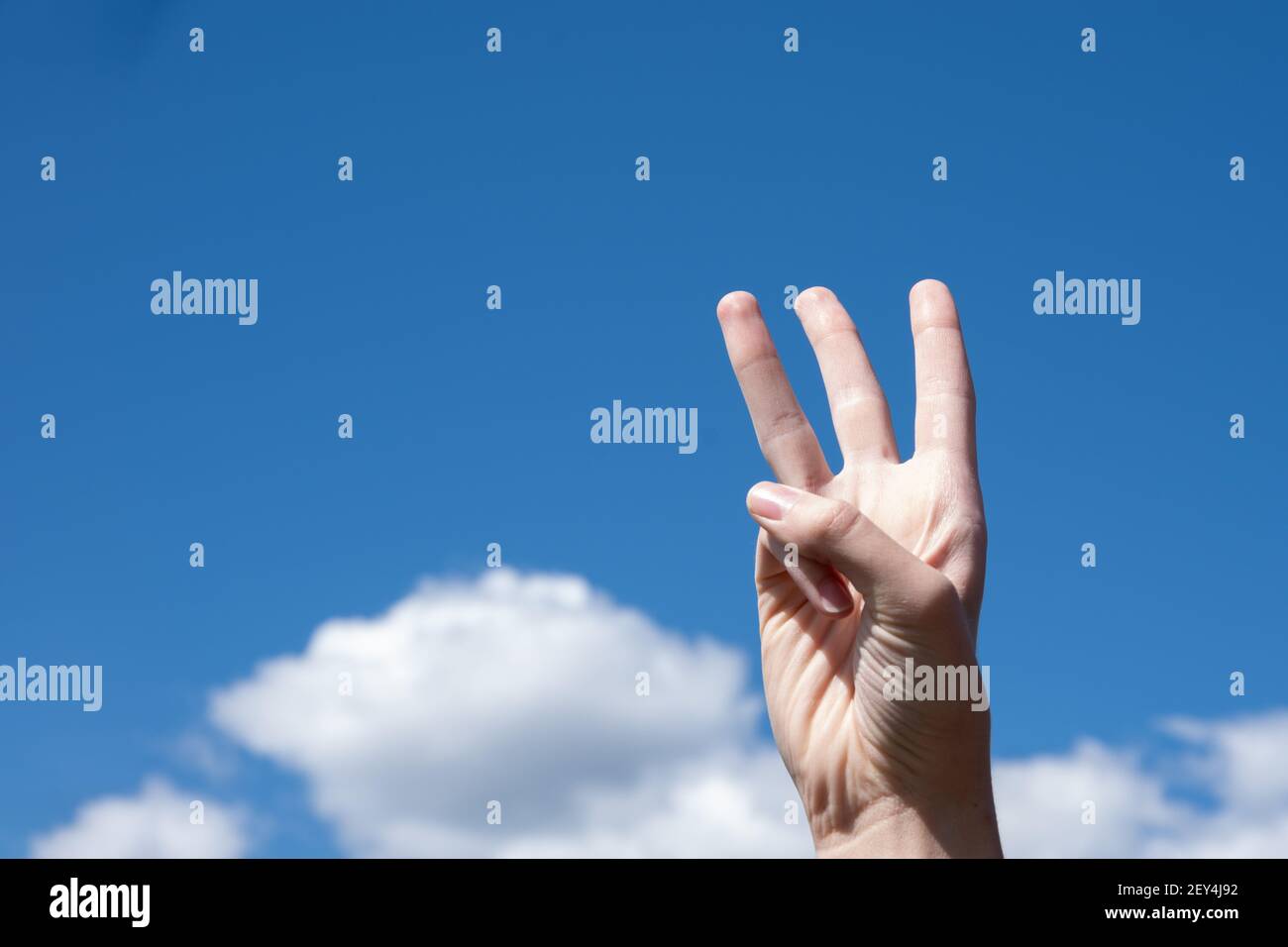 Close-up gesture of a woman's hand showing three fingers isolated on a background of blue sky with clouds, symbol sign language number three. Stock Photo