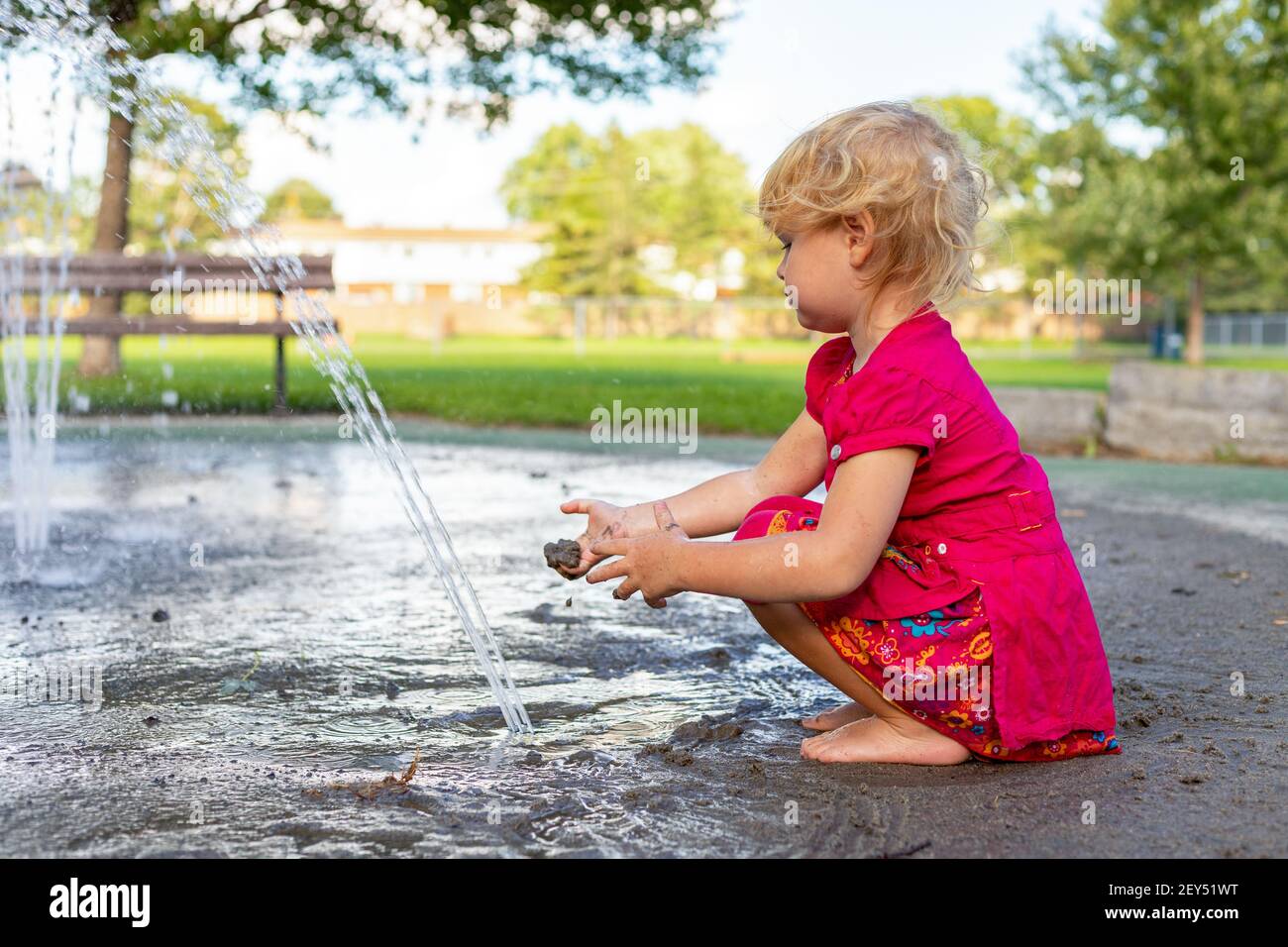 Little girl playing with splash pad water and dirt in the park on sunny day in summer. Stock Photo