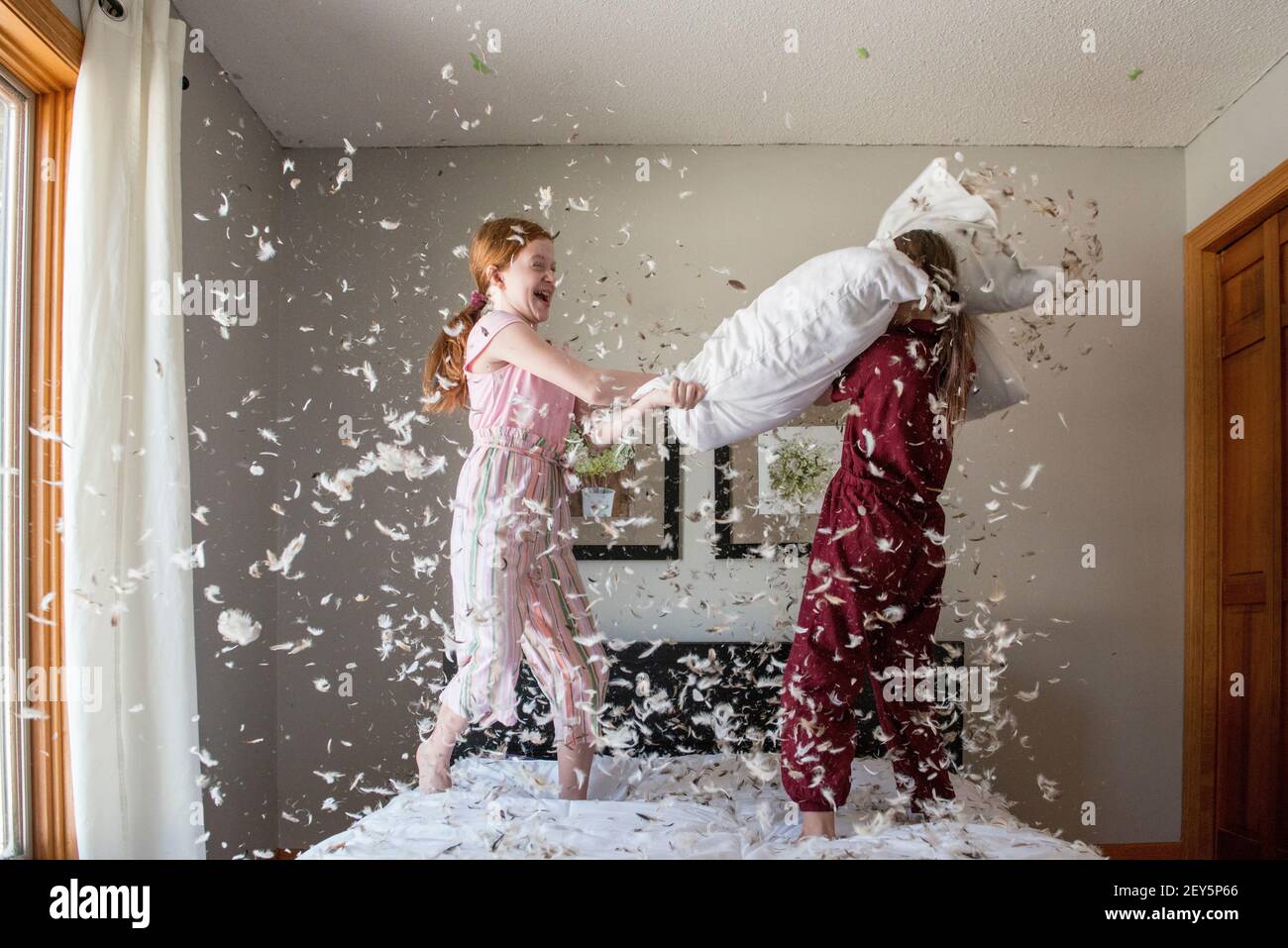 Two happy young girls having a feather pillow fight on the bed. Stock Photo