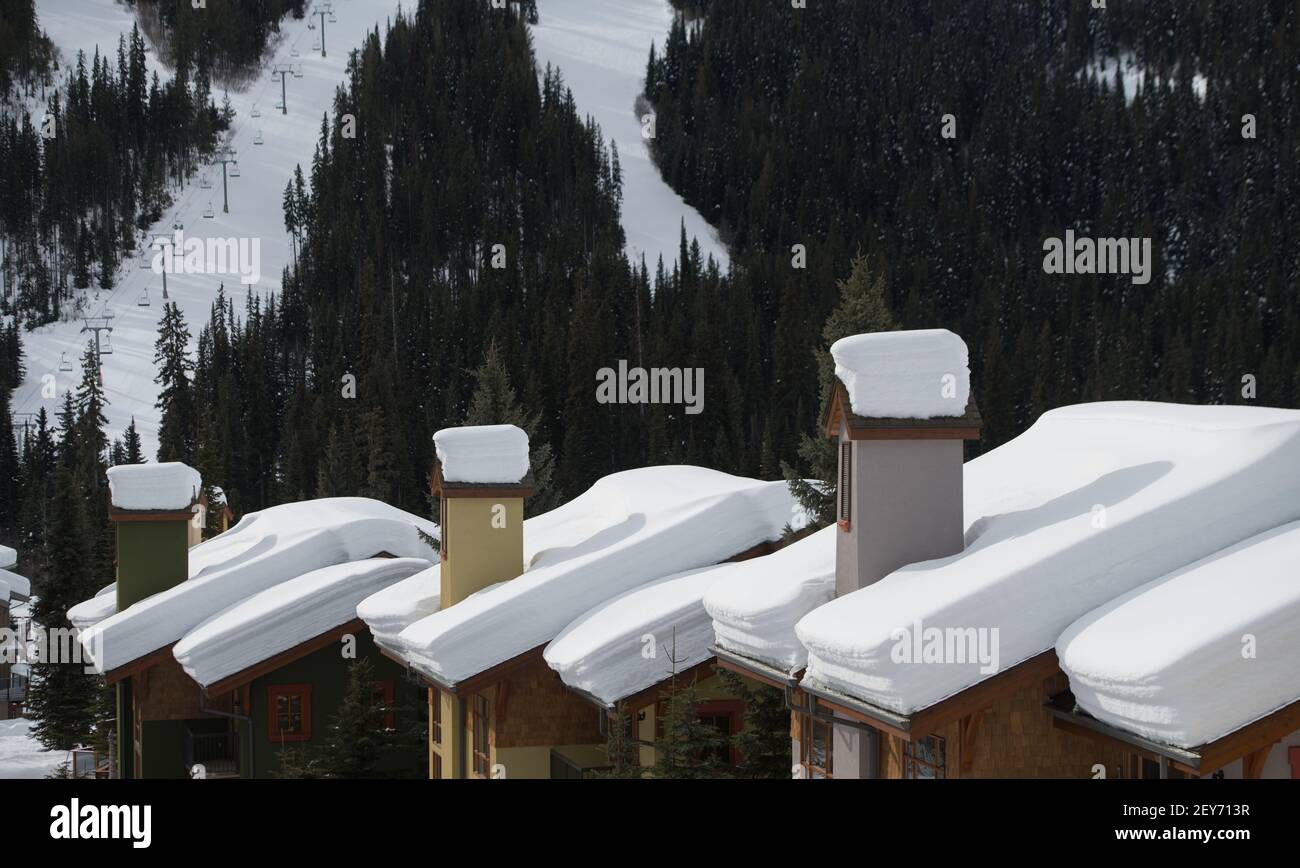 fresh snow and layers of snow sitting on roof of ski chalets with tall chimneys ski runs in background ski lift or chair lift in background horizontal Stock Photo