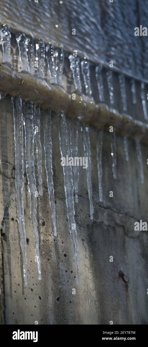 frozen pointy icicles hanging from industrial metal pipe on grungy water damaged concrete building ice shadows on exterior concrete wall in background Stock Photo