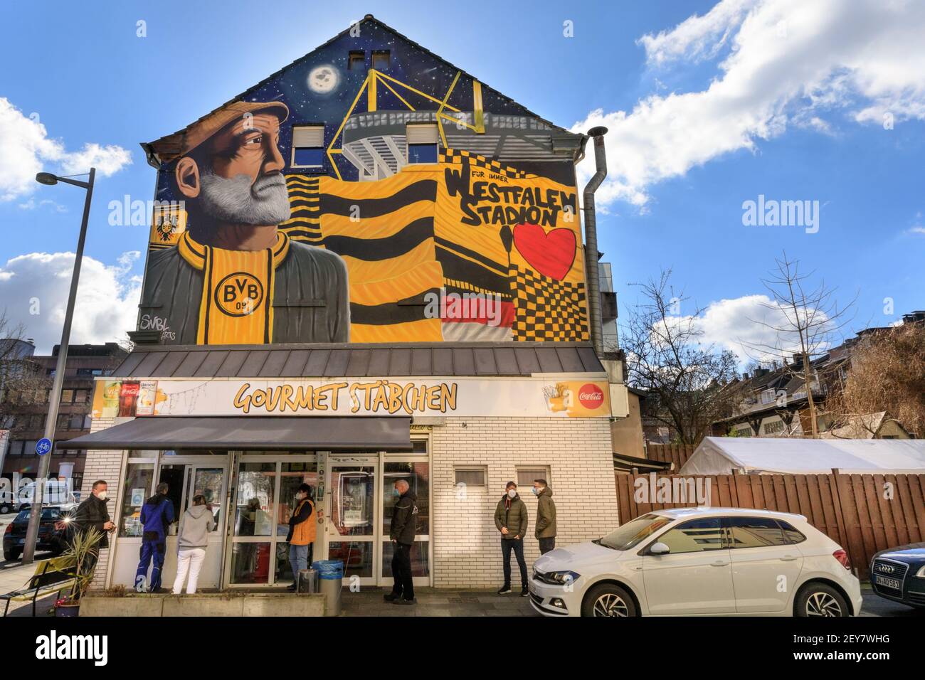 Dortmund, NRW, Germany. 05th Mar, 2021. People patiently queue for their food outside the Gourmet Stäbchen take away, decorated with a mural of Borussia Dortmund football club's stadium, colours and a fan. The club plays an important part in the city's sporting life and cultural identity. North Rhine-Westphalia saw beautiful but cold sunshine today in most areas. Credit: Imageplotter/Alamy Live News Stock Photo