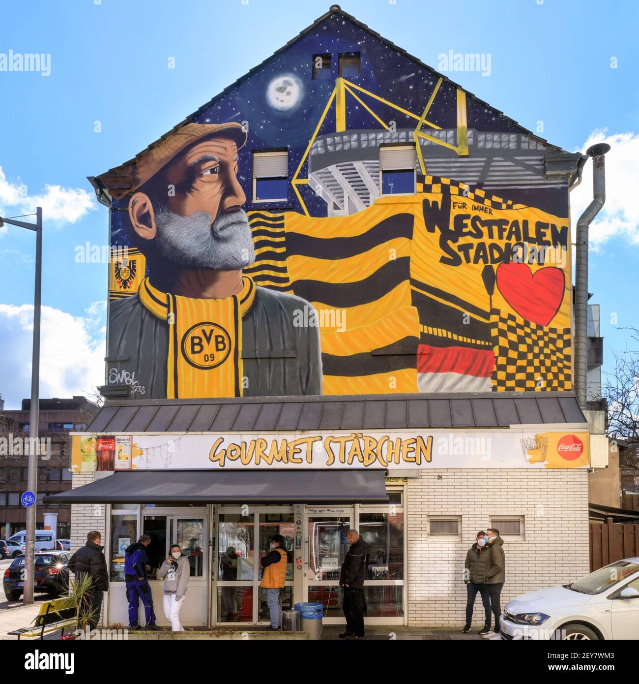 Dortmund, NRW, Germany. 05th Mar, 2021. People patiently queue for their food outside the Gourmet Stäbchen take away, decorated with a mural of Borussia Dortmund football club's stadium, colours and a fan. The club plays an important part in the city's sporting life and cultural identity. North Rhine-Westphalia saw beautiful but cold sunshine today in most areas. Credit: Imageplotter/Alamy Live News Stock Photo