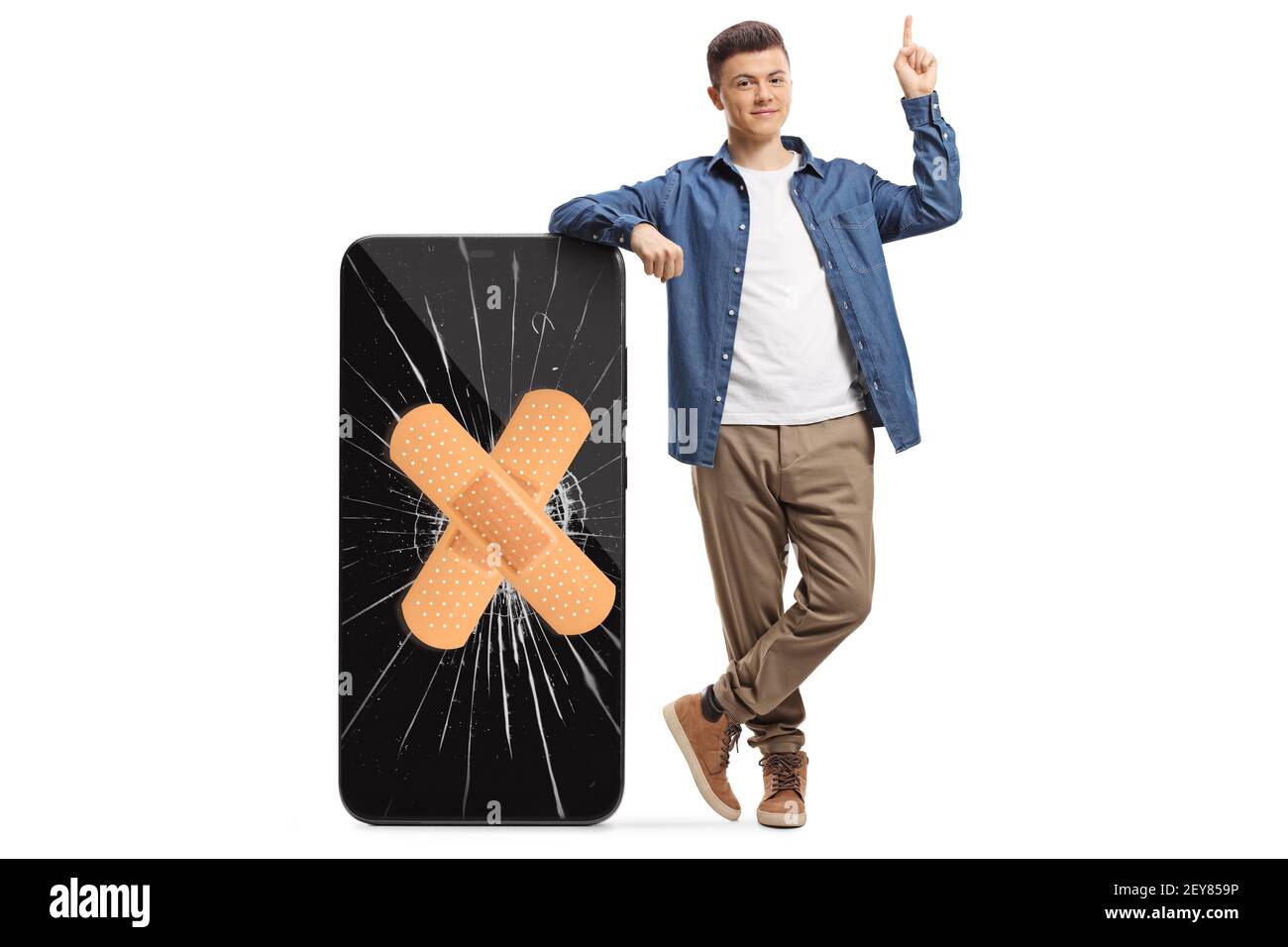 Full length portrait of a guy leaning on a big smartphone with cracked screen and bandage and pointing above isolated on white background Stock Photo