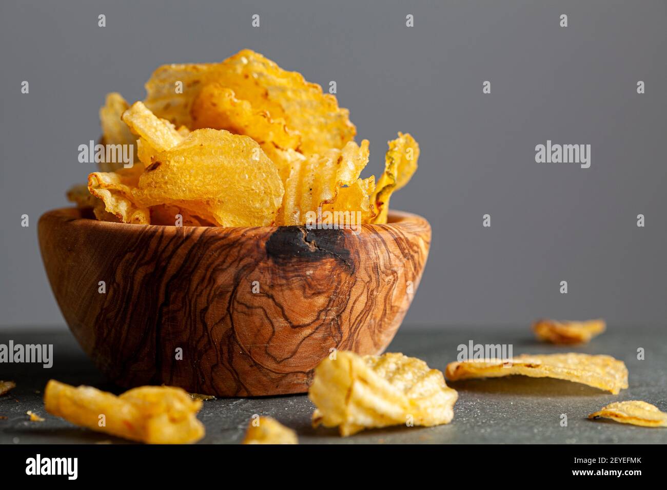A wooden bowl of kettle cooked potato chips with crunchy ruffled texture. Some are spilt on the dark background. A moody pub, bar feeling or kitchen t Stock Photo