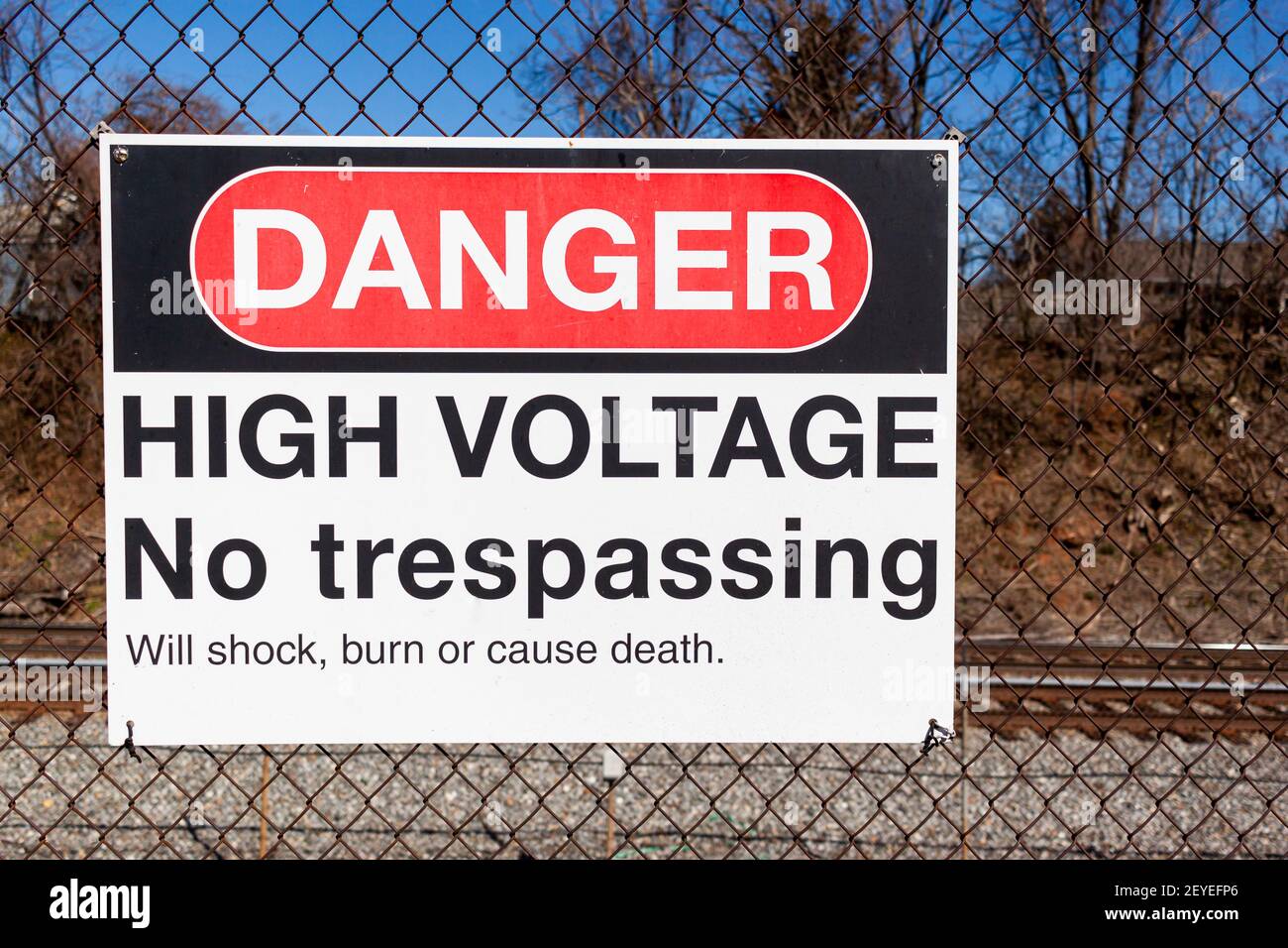 Danger, high voltage, no trespassing sign on a fence in front of a rail road. It lists possible health hazards including shock, trauma and death under Stock Photo