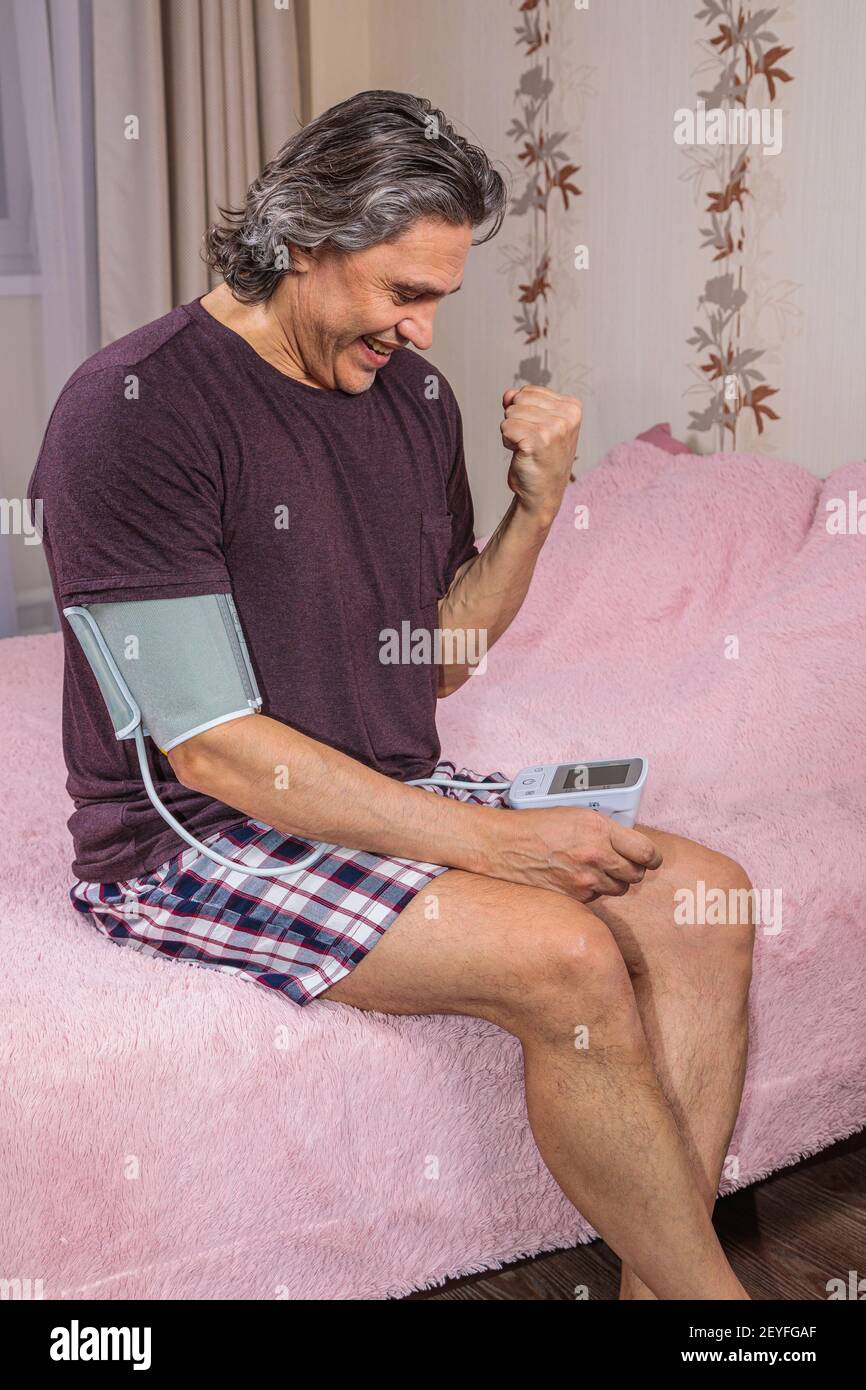 A 50-year-old man measures his blood pressure with a blood pressure monitor at home, sitting on his bed in his home clothes. Surprised by the instrume Stock Photo
