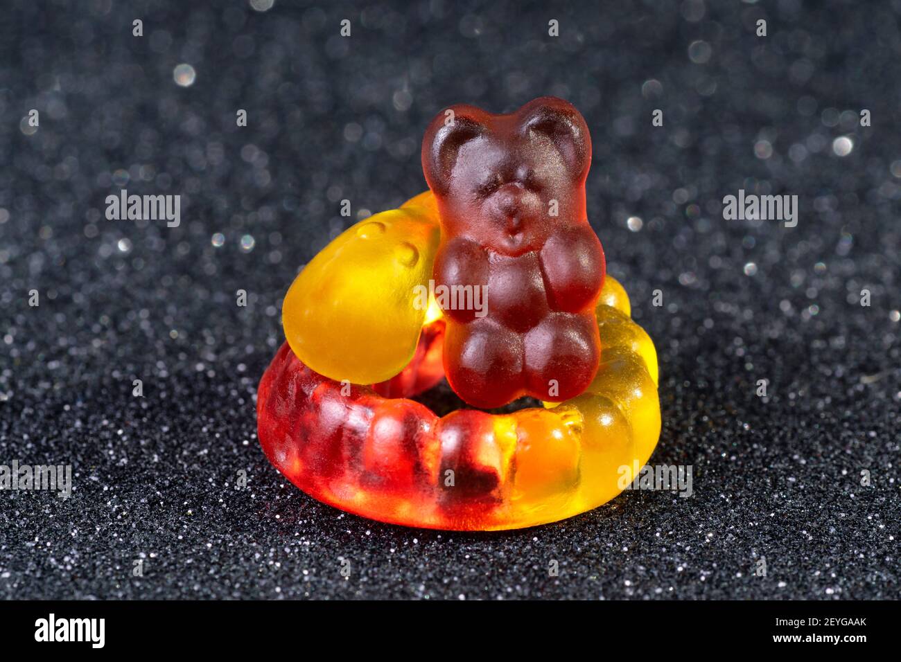Close-up of a purple gummy bear sitting on a curled yellow-red gummy worm on a glittering blurred background. Stock Photo