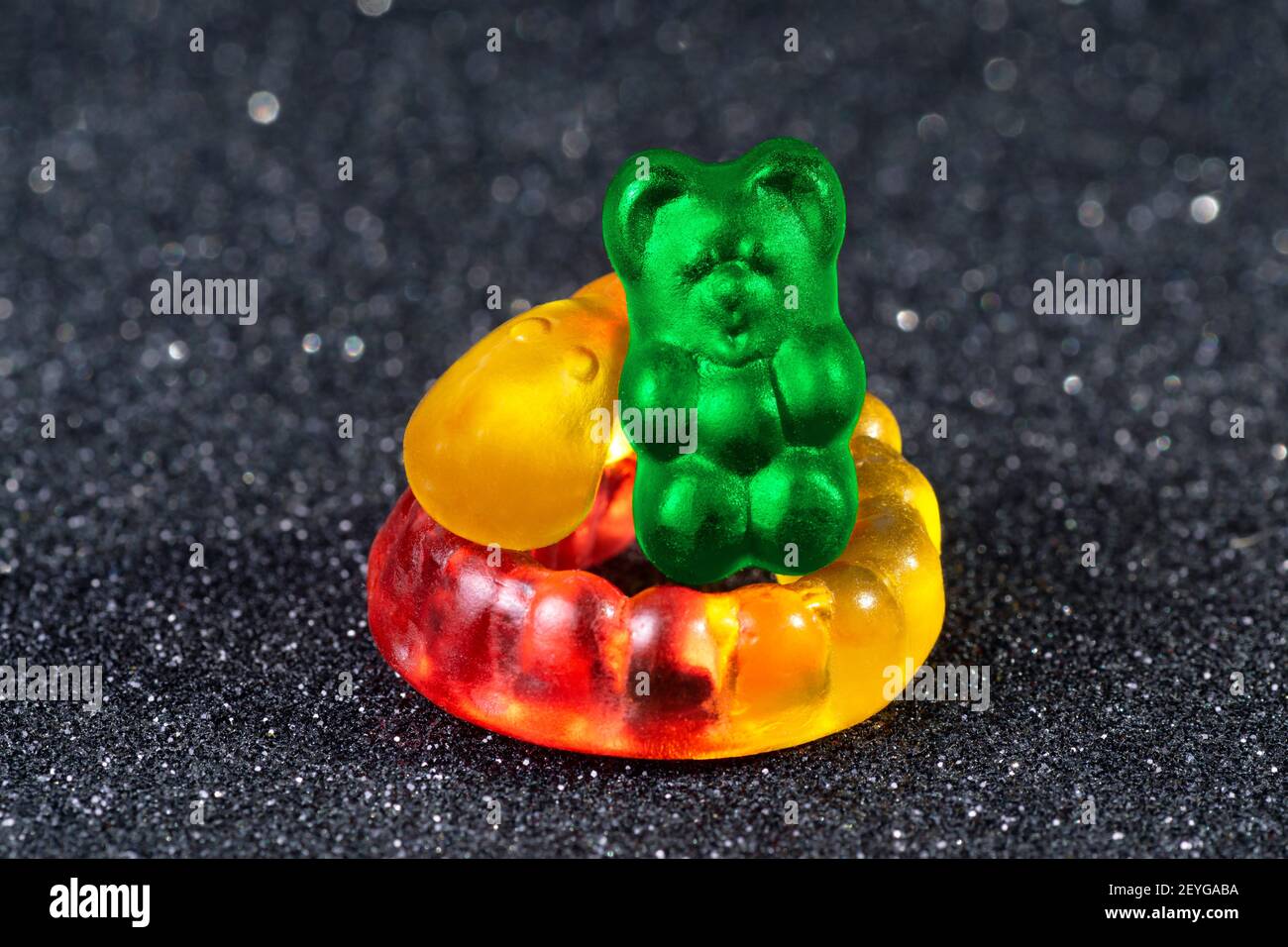 Close-up of a green gummy bear sitting on a curled yellow-red gummy worm on a glittering blurred background. Stock Photo