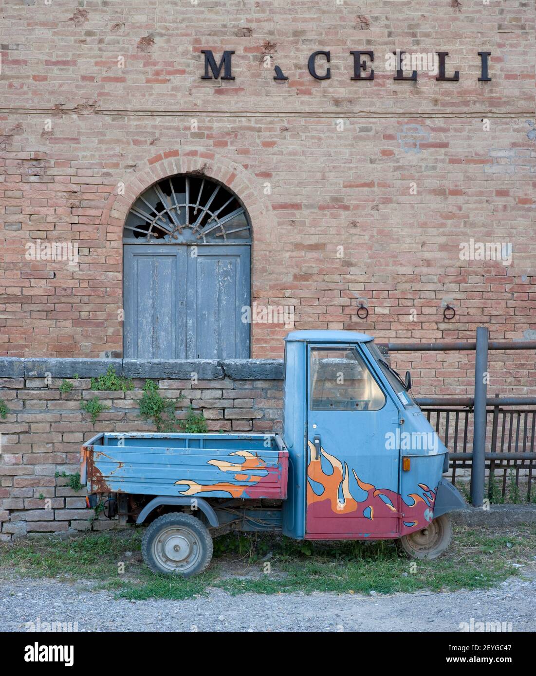 old Piaggio Ape (bee) with flames painted on its side in front of old factory Tuscany, near Buonconvento,Italy, alter Piaggio Ape (Biene) vor Fabrik Stock Photo