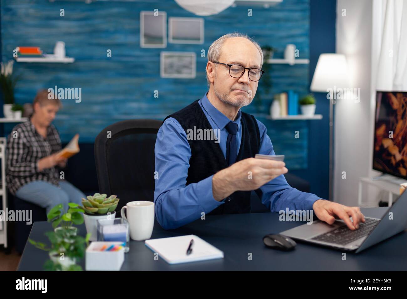 Senior man reading cvv conde on credit card wearing glasses. Elderly man checking online banking to make shppping payment looking at laptop while wife is reading a book sitting on sofa. Stock Photo