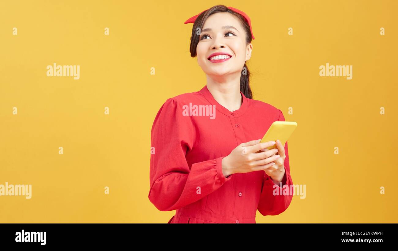 Portrait of a smiling young casual asian woman looking at mobile phone isolated over yellow background Stock Photo