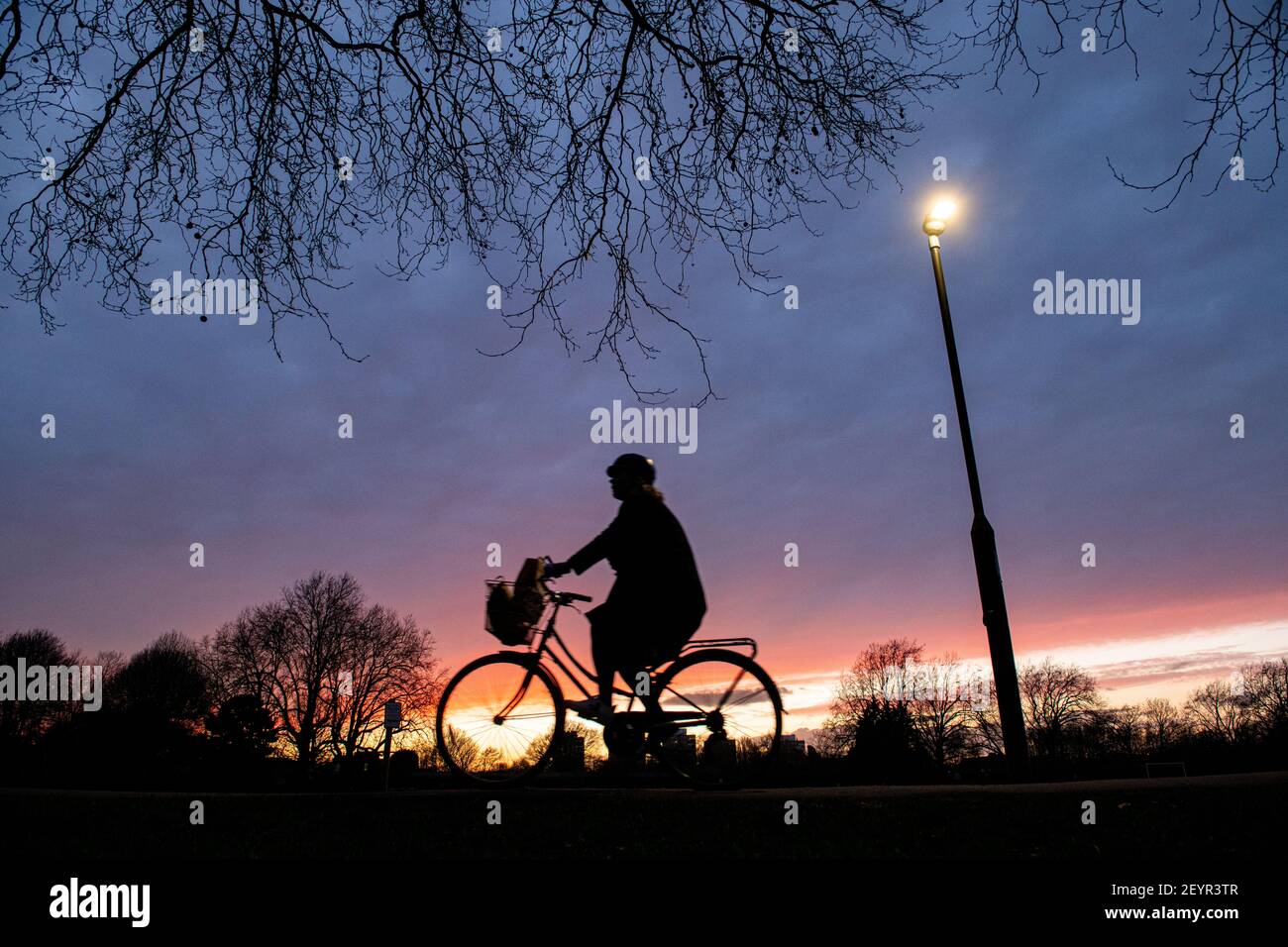 A cyclist rides through a London park at sunset under  street light Stock Photo
