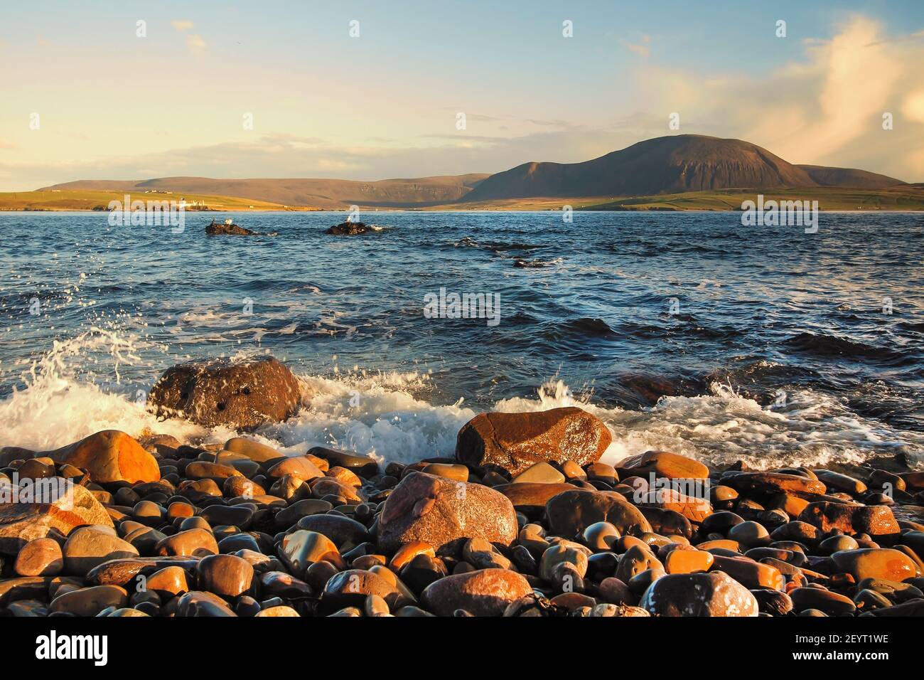 Shoreline with round stones and big hill in background on summer sunny afternoon on Orkney islands in northern Scotland Stock Photo