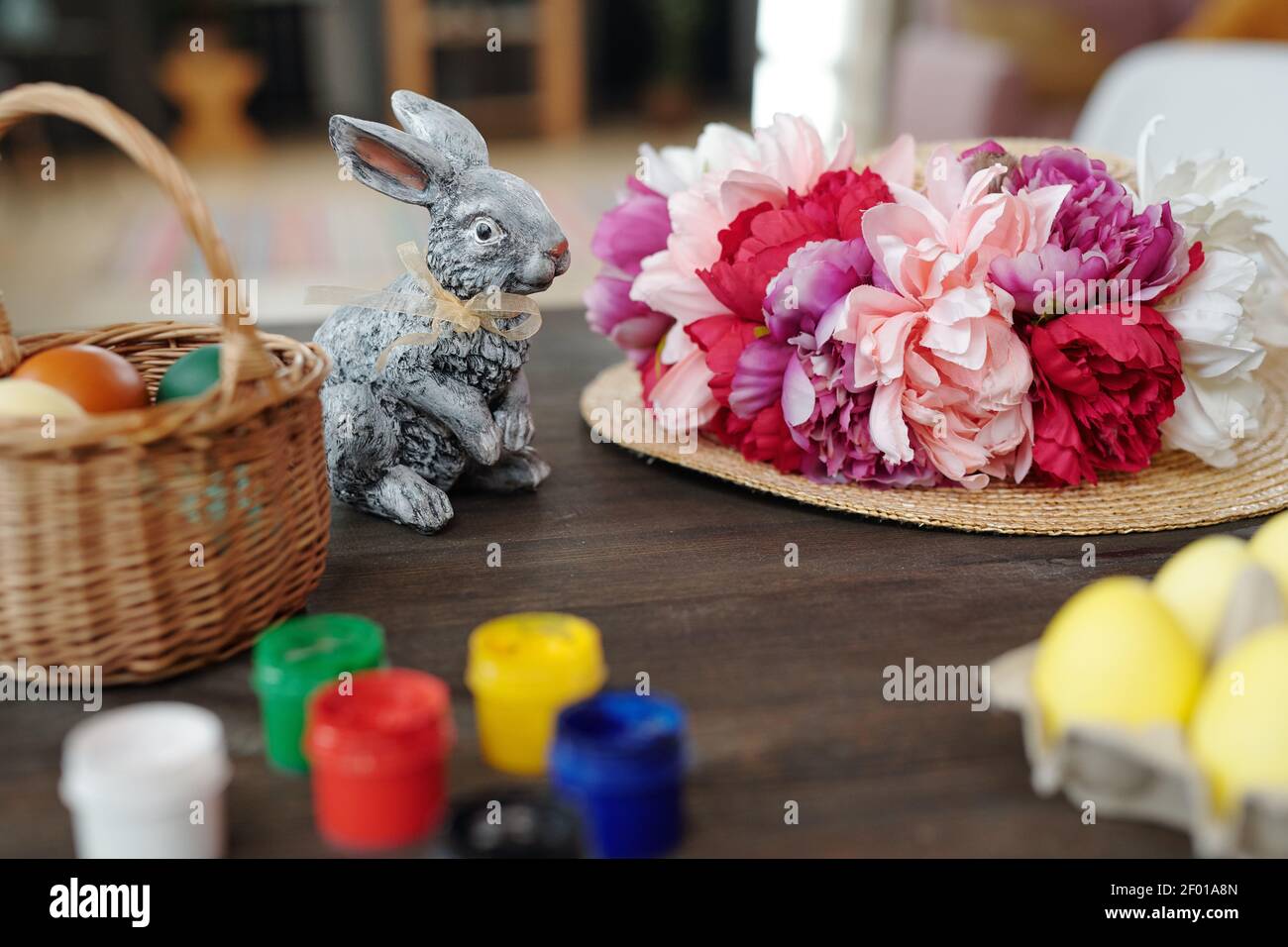 Grey toy rabbit, basket with painted Easter eggs and hat with handmade white, pink and crimson flowers on wooden table in front of camera Stock Photo
