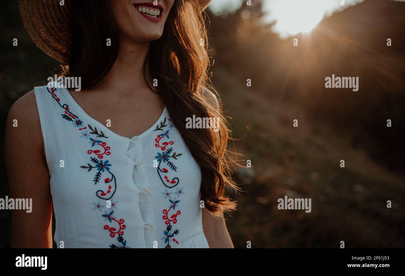 The midsection of a young smiling woman in light summer clothes wearing a hat letting her long hair shine under the sunset rays Stock Photo